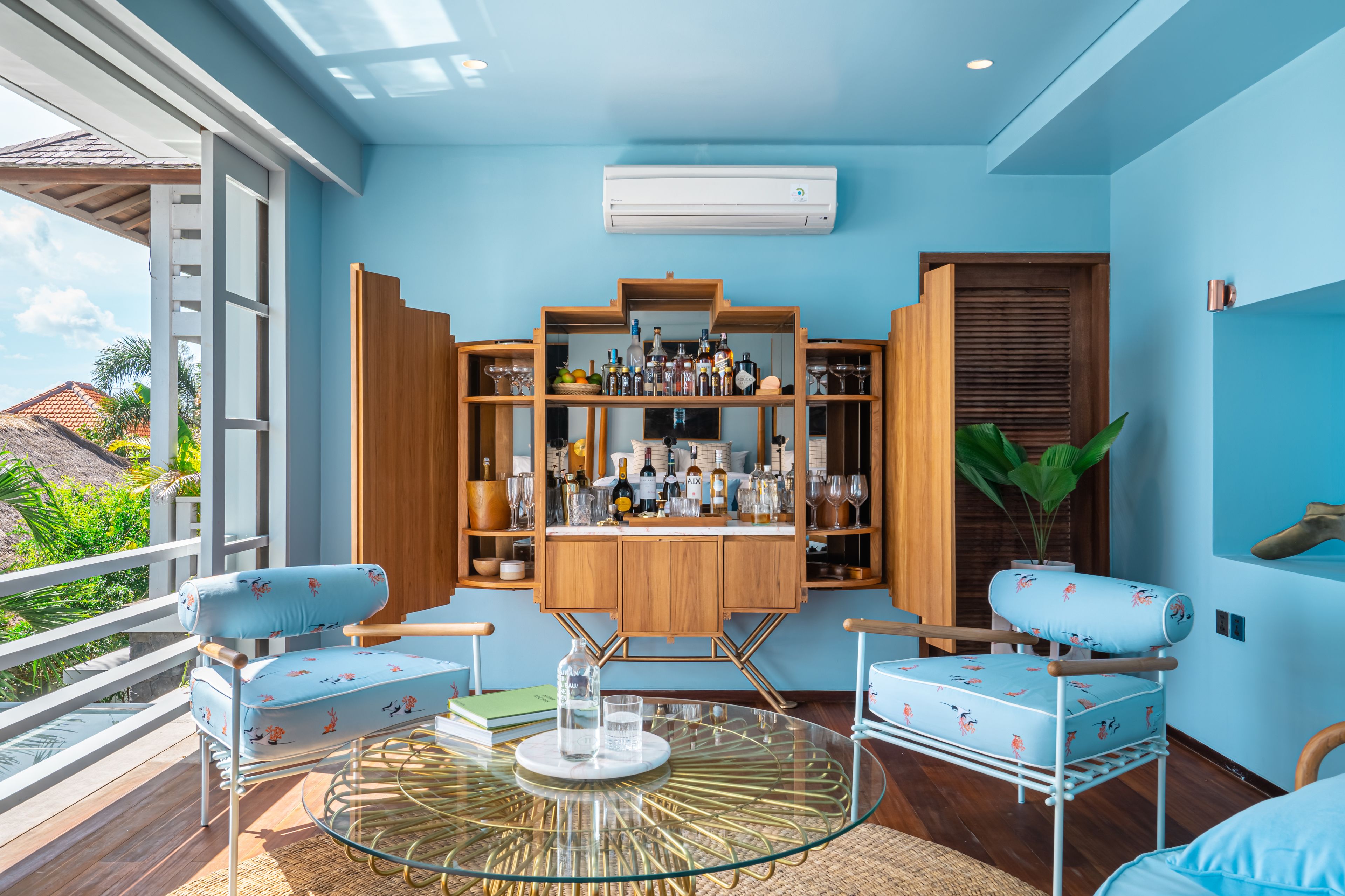 A room with a blue wall, a wooden cabinet with bottles, two blue chairs, a glass table, and a view of greenery outside.