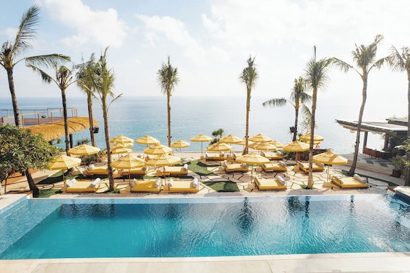 A poolside area with lounge chairs and umbrellas, palm trees, and a view of the ocean. - Ulu Cliffhouse - Adult Only (Pecatu)