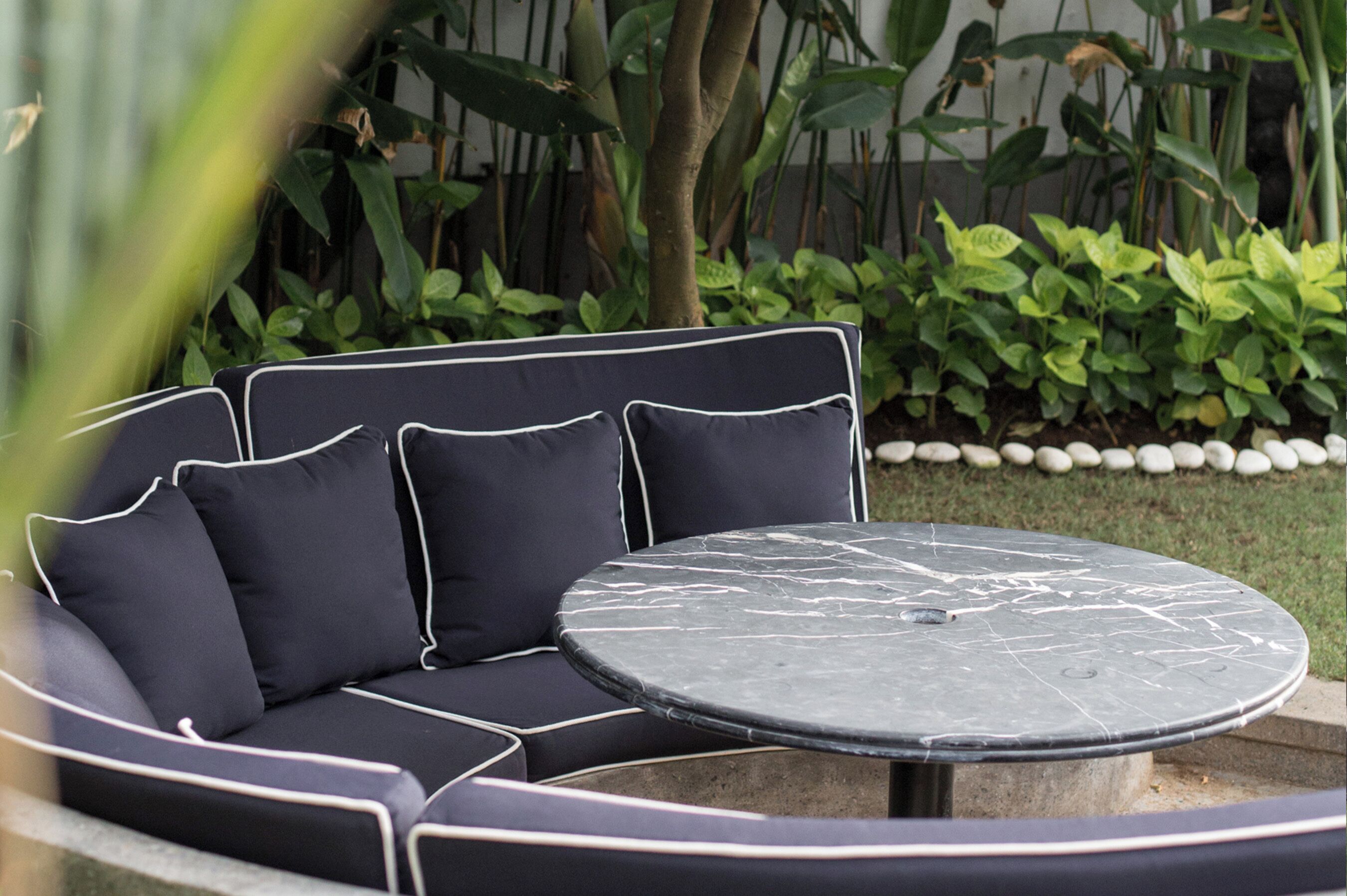 A black outdoor sofa with white piping, a round marble table, and a garden setting with plants and a stone border.