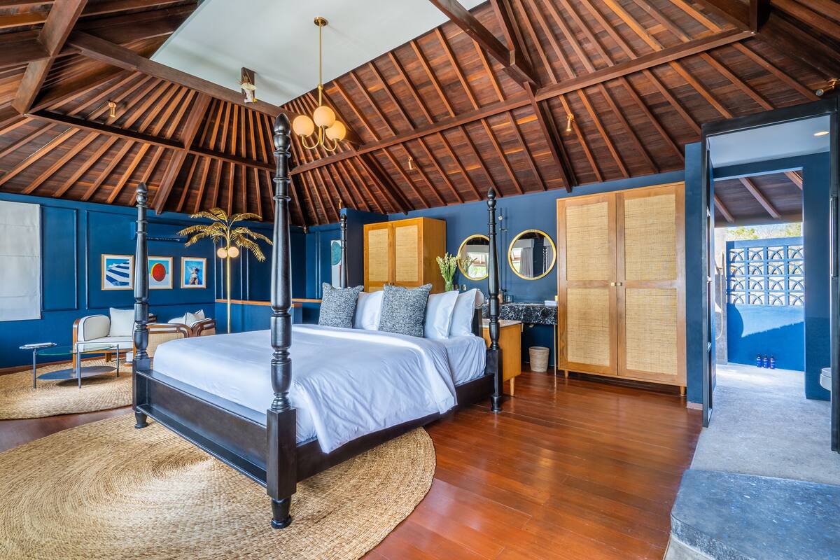A spacious bedroom with a four-poster bed, wooden ceiling beams, and a large window.