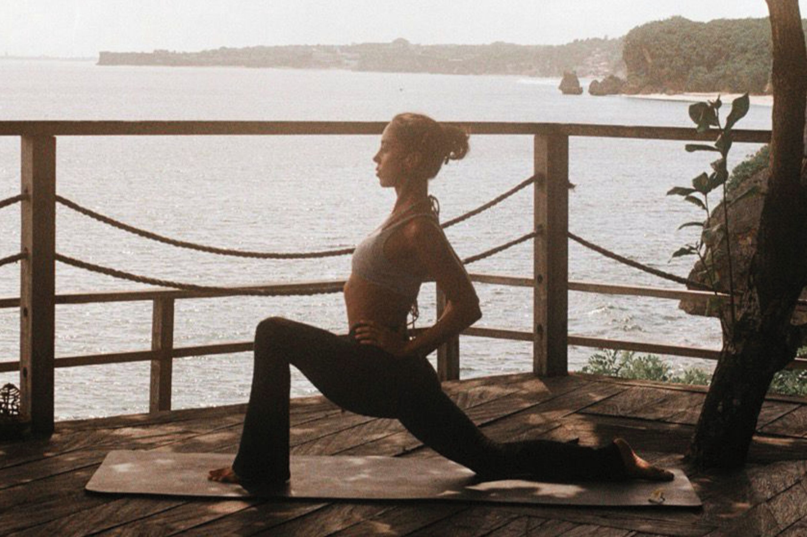 A person performing a yoga pose on a mat by a wooden deck with a view of the sea.