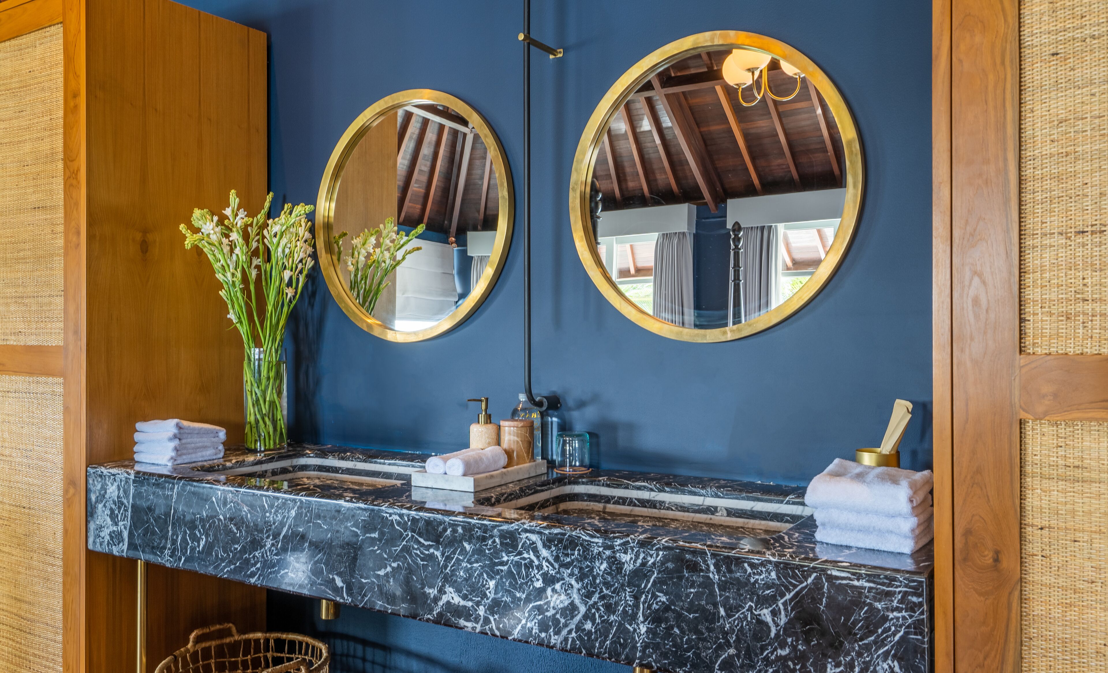 A bathroom with a marble vanity, two round mirrors, a vase with flowers, and toiletries.