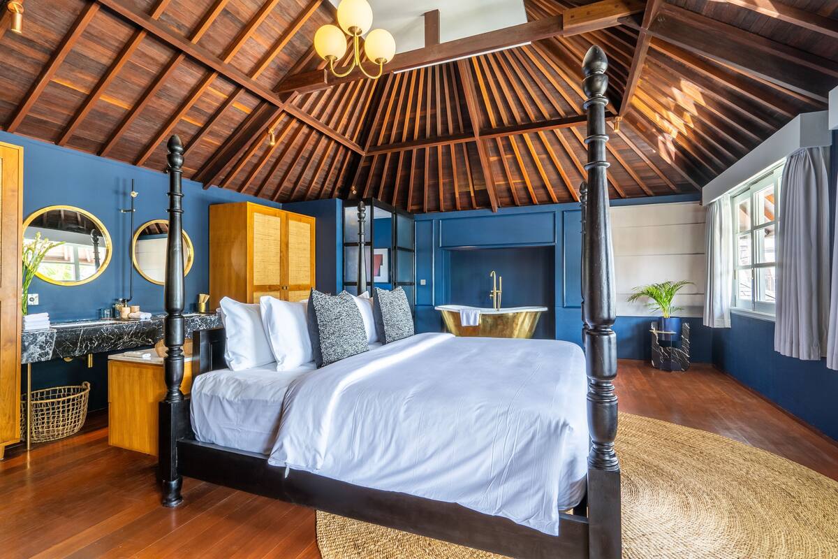 A four-poster bed with a dark finish, a wooden ceiling with exposed beams, a round mirror, a small round table, and a window with curtains.