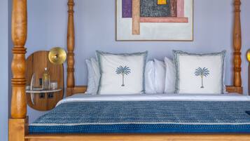 A wooden four-poster bed with a blue blanket, two pillows with palm tree designs, and a framed abstract painting on the wall.