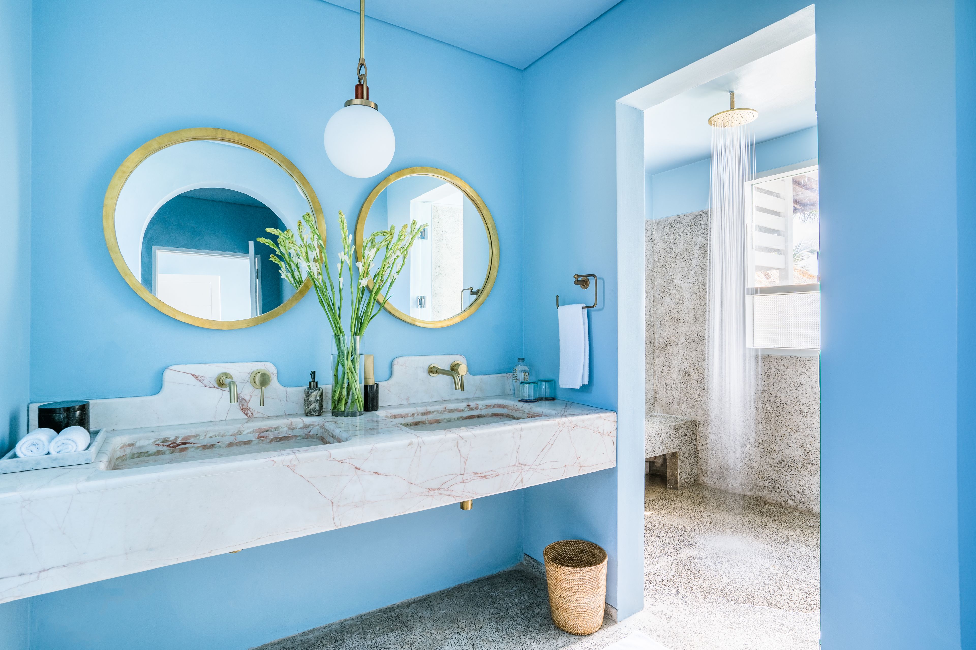 Un baño con una pared azul, dos espejos redondos con marcos dorados, un lavamanos de mármol blanco y una zona de ducha.