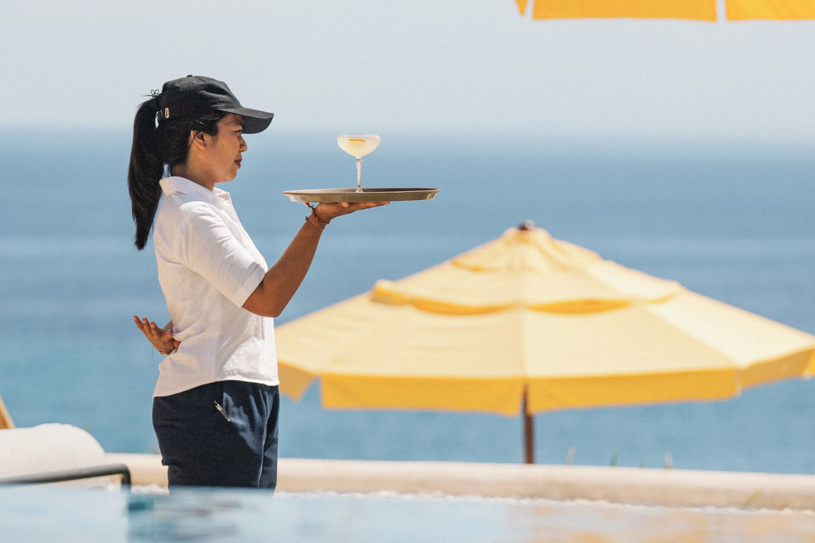A person holding a tray with a drink near a poolside with umbrellas.