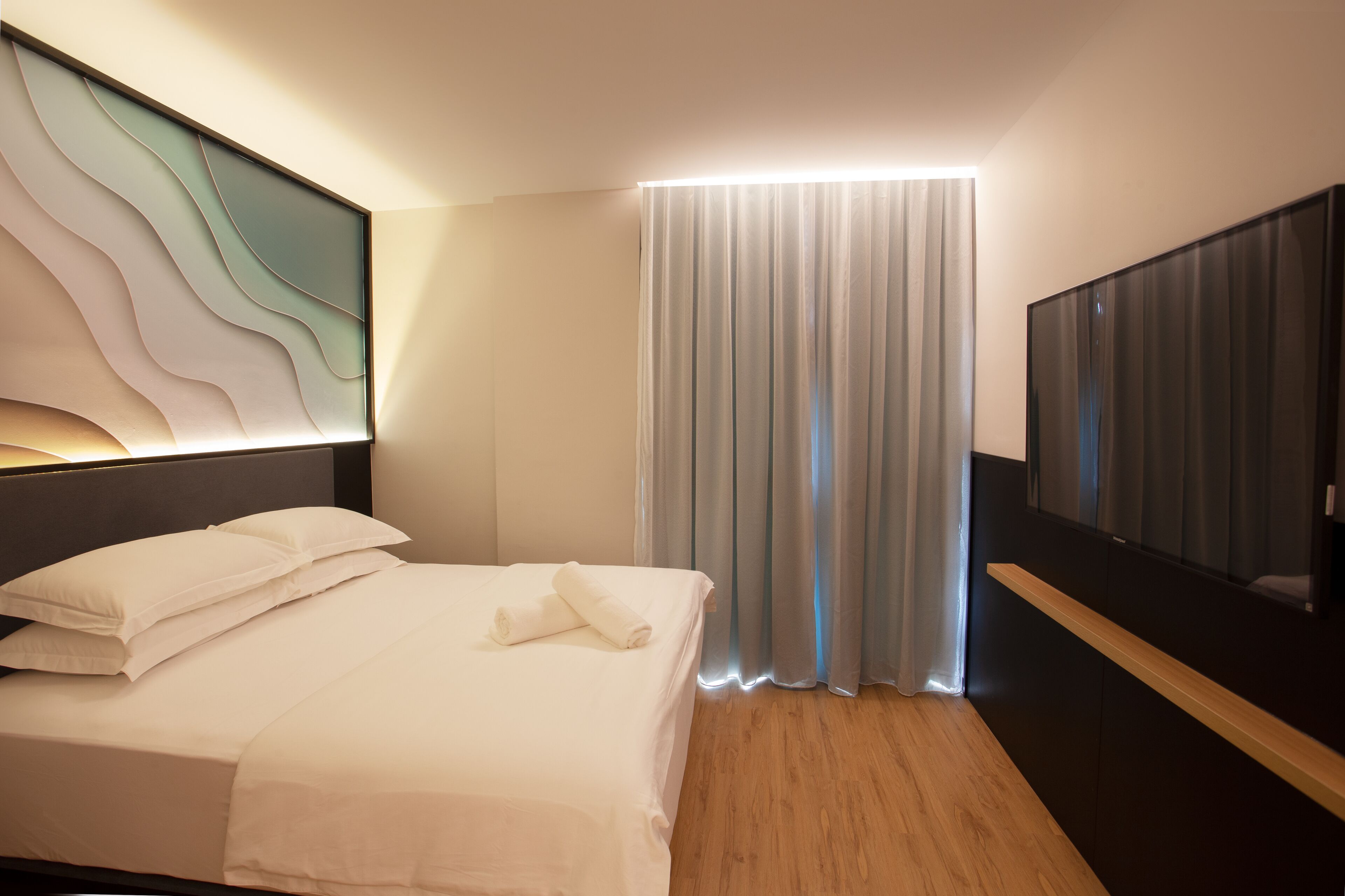 A modern hotel room with a neatly made bed, a large flat-screen TV, and a large window with curtains.