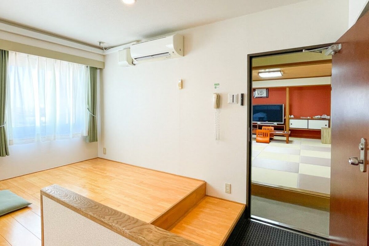 A room with a wooden floor, a bed, a desk, a chair, a window with curtains, an air conditioning unit, and a door leading to another room.