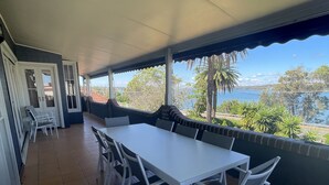 A covered outdoor dining area with a white table and chairs, a bench, and a view of the sea and trees. - SPEERS POINT - Water Views, Pool, Sleeps 12, 8 Beds, Great for Families/groups (Speers Point)