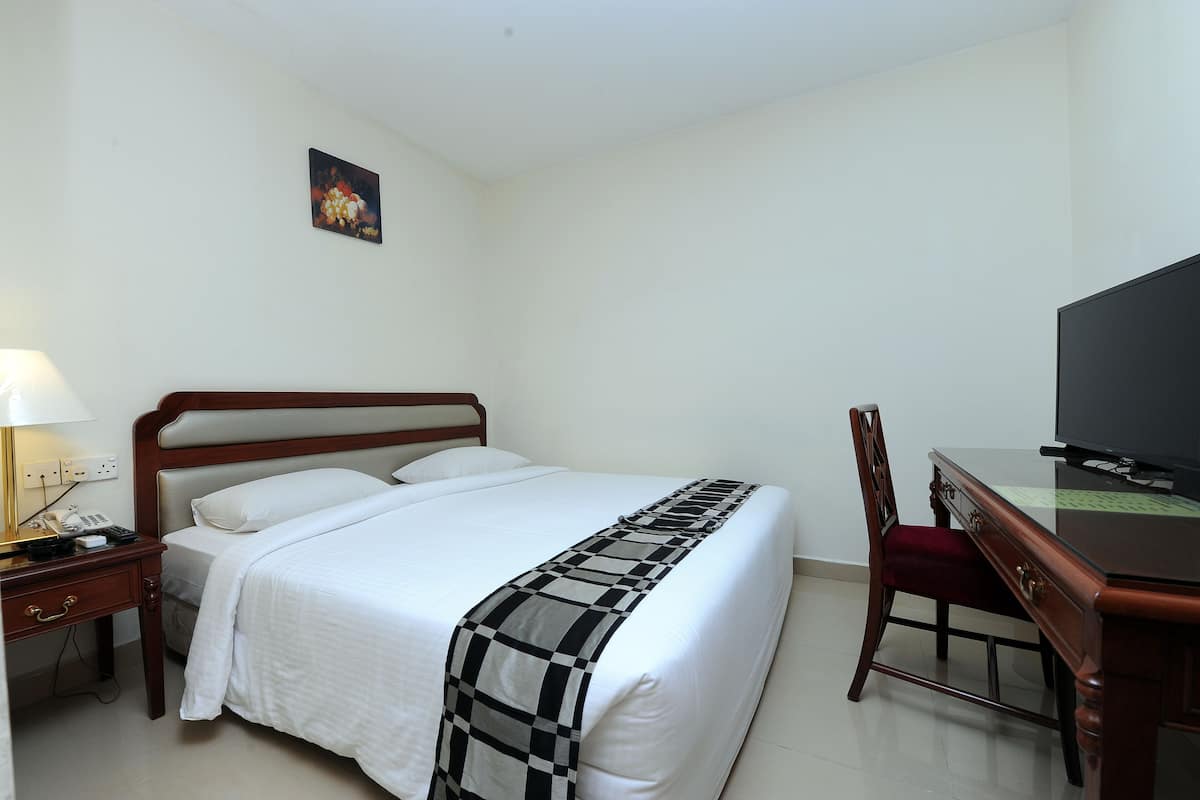 A neatly made bed with a checkered blanket, a wooden nightstand with a lamp, a desk with a television, and a chair.