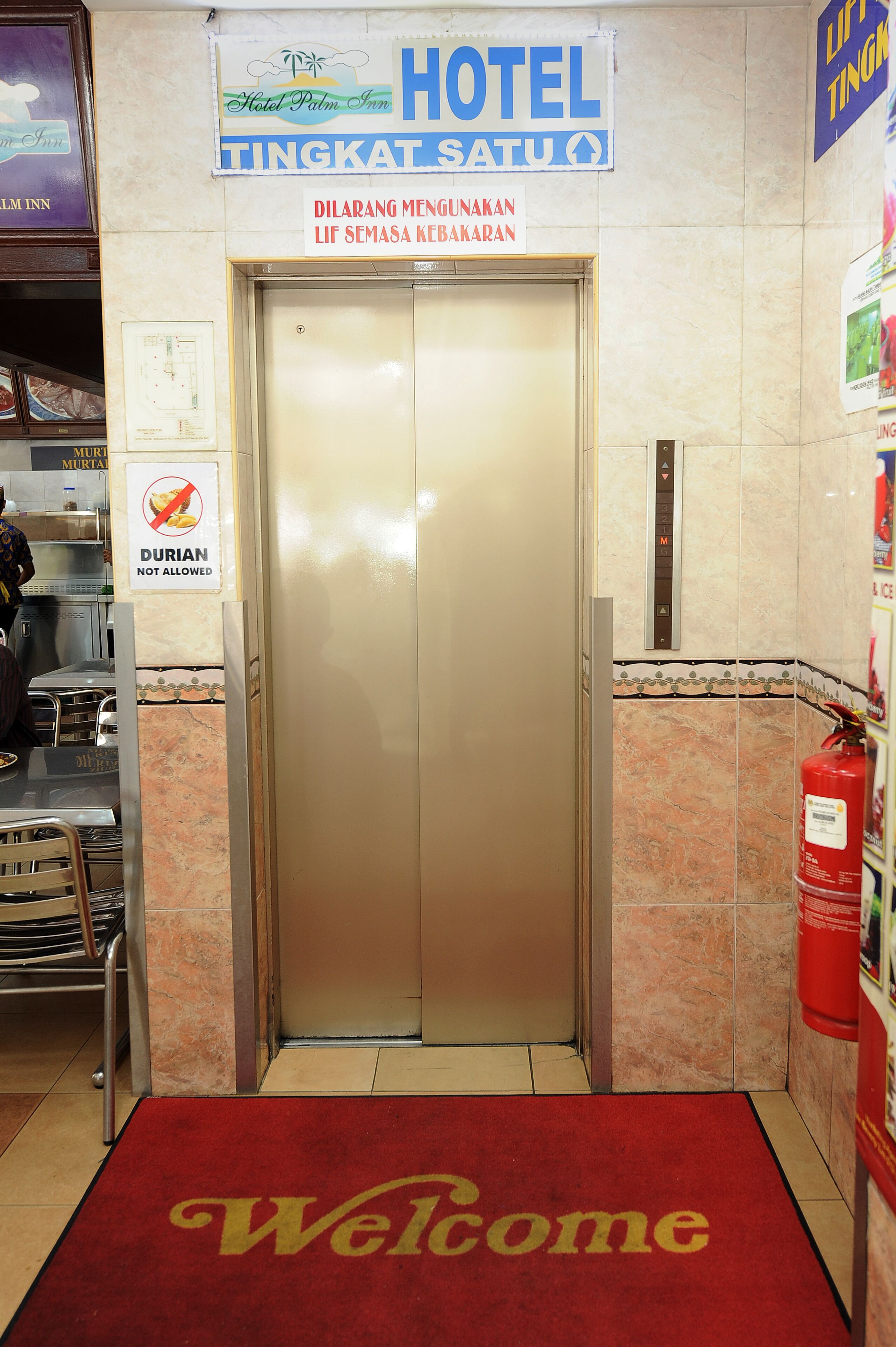 Een hotelentree met een welkomstmat, een lift en een bord waarop staat 'DILARANG MENGUNAKAN LIF SEMASA KEBAKARAN'.