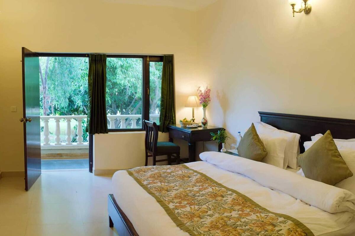 A hotel room with a bed, a desk, a chair, a lamp, and a view of greenery through the open door.