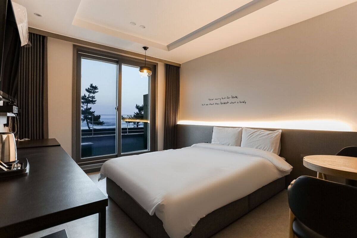 A modern hotel room with a large bed, a desk, a chair, and a view of the sea through a large window.