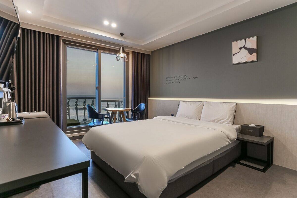 A modern hotel room with a large bed, a desk, and a view of the sea through the window.