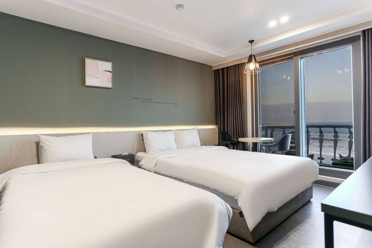 A hotel room with two beds, a small table, a chair, and a view of the sea through large windows.