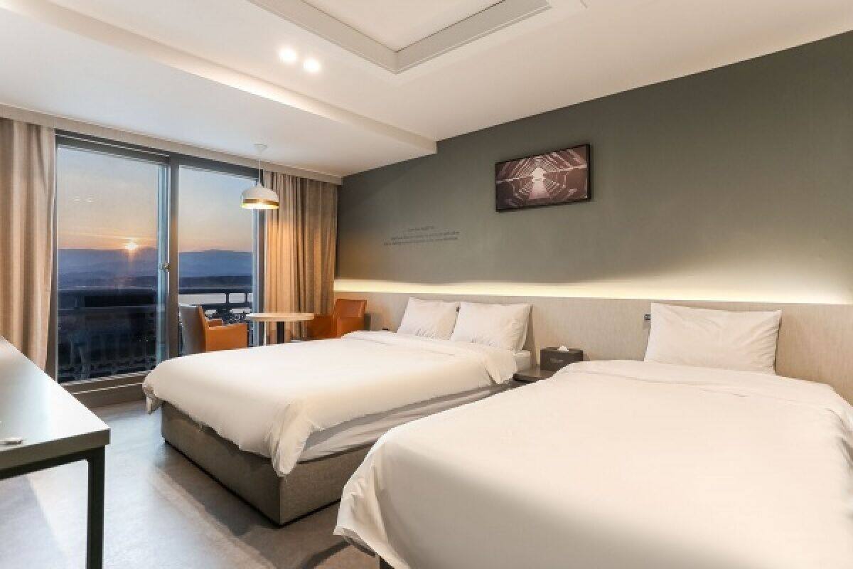 A hotel room with two beds, a desk, and a large window with a view of the sunset.