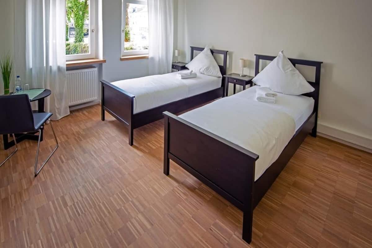 Une chambre d’hôtel avec deux lits simples, une petite table, une chaise et une fenêtre avec des rideaux.