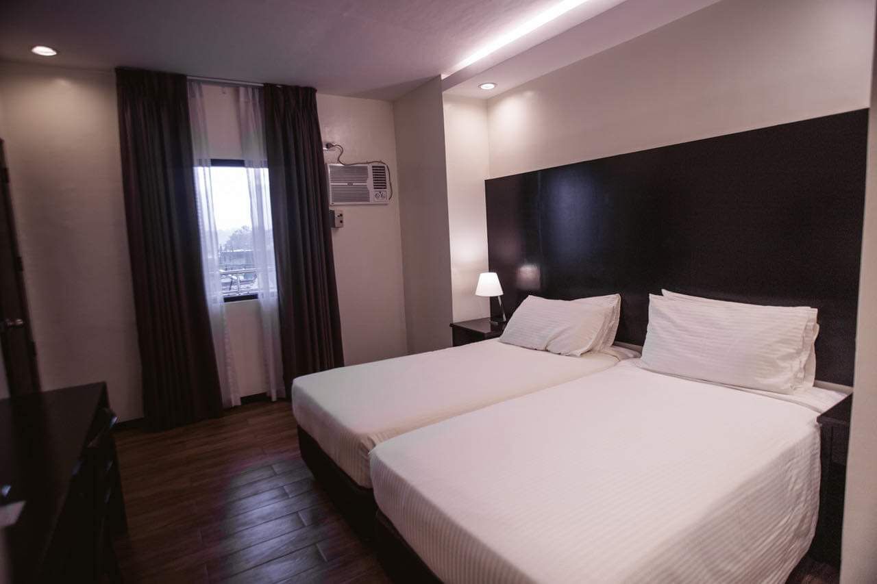A hotel room with two beds, a desk, a window with curtains, an air conditioning unit, and a nightstand with a lamp.