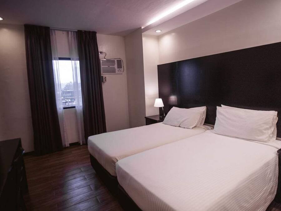 A hotel room with two beds, a desk, a window with curtains, an air conditioning unit, and a nightstand with a lamp.