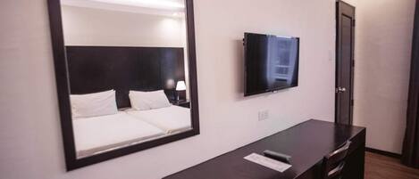 Habitación de hotel con un espejo grande, un televisor de pantalla plana en la pared, un escritorio de madera oscura y una cama con ropa blanca.