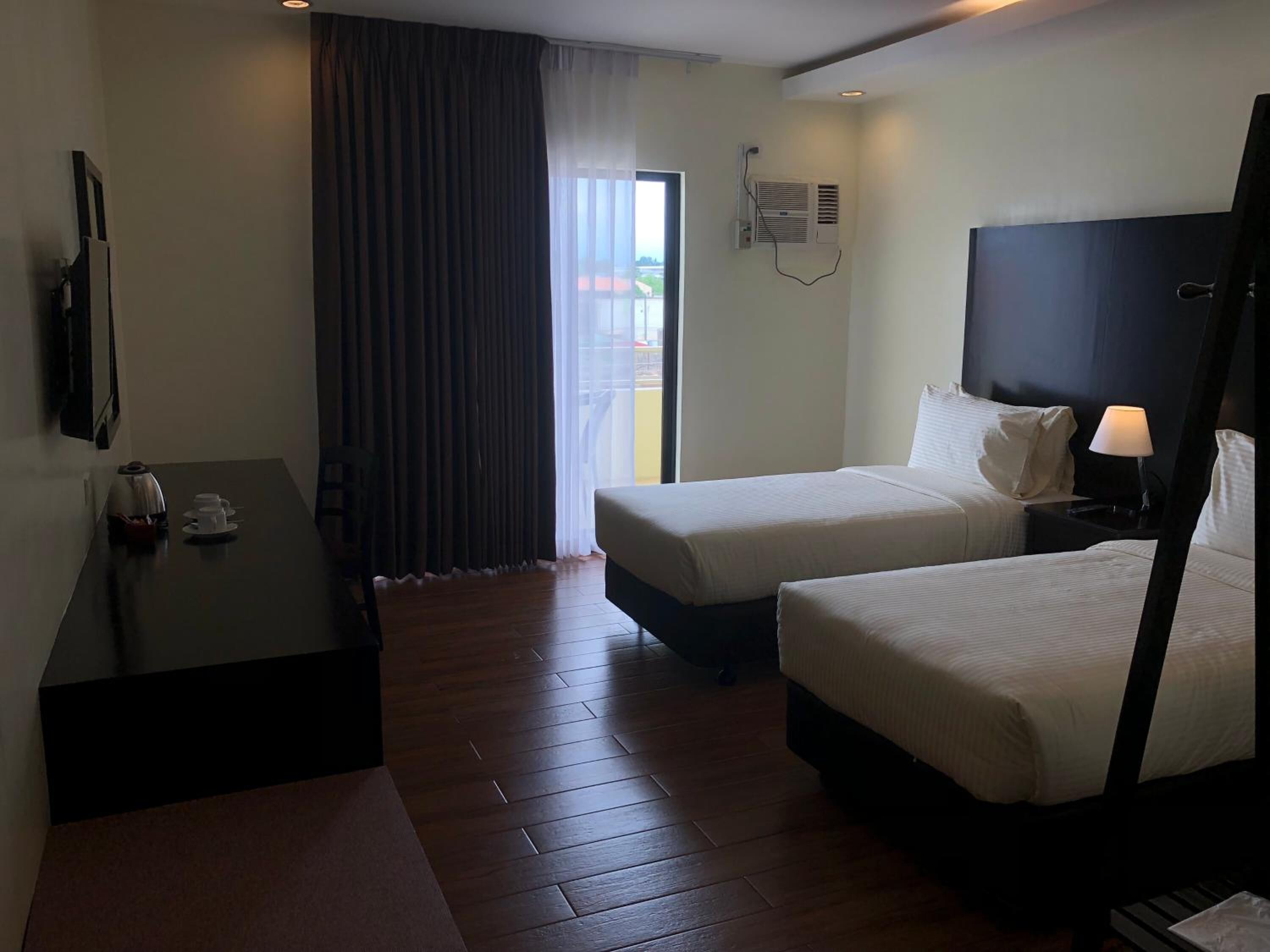 A hotel room with two beds, a small table with a kettle and cups, a TV on the wall, and a window with curtains.