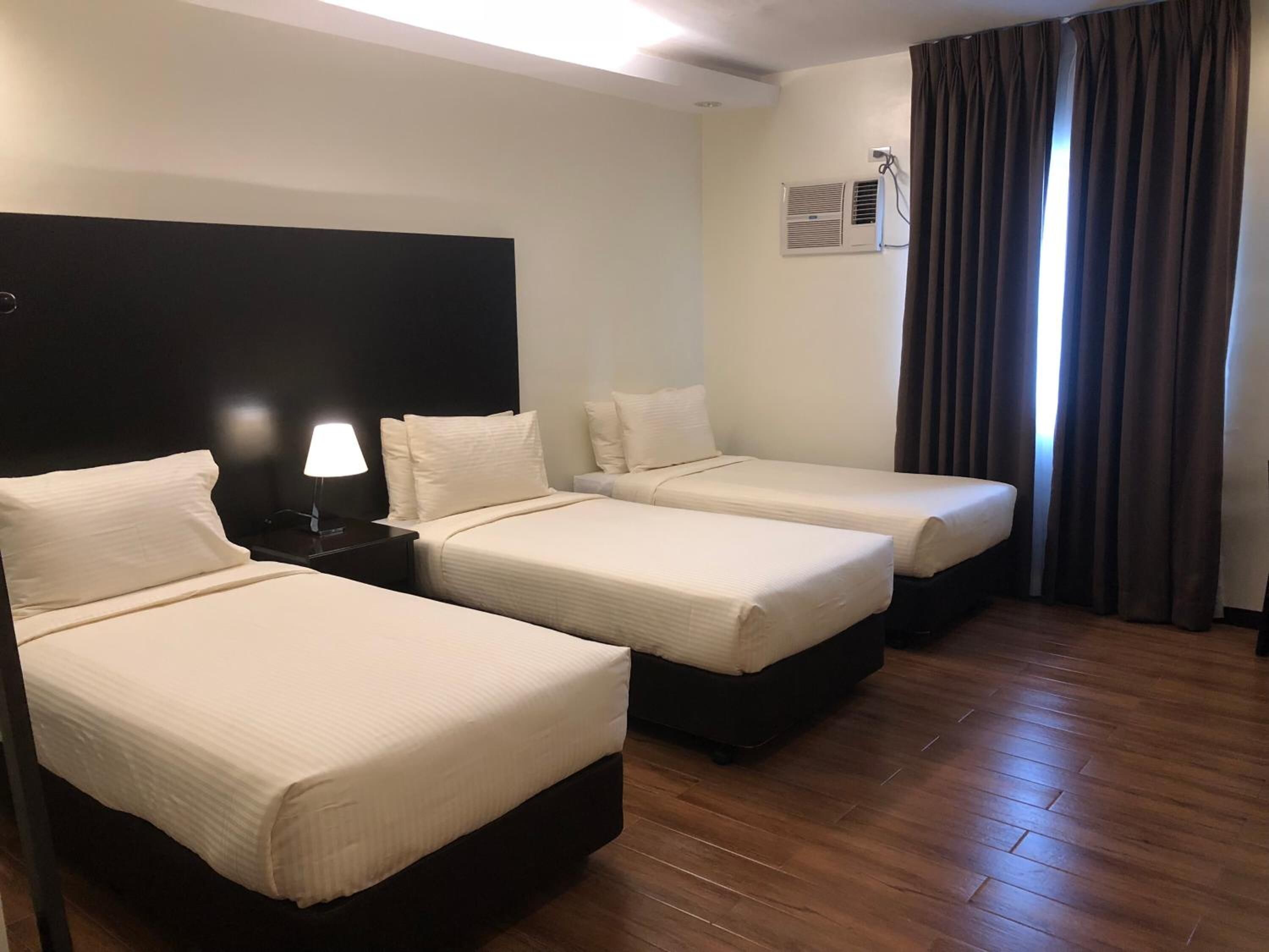 Habitación de hotel con dos camas individuales, una mesita de noche con lámpara, aire acondicionado instalado en la pared y una ventana con cortinas.
