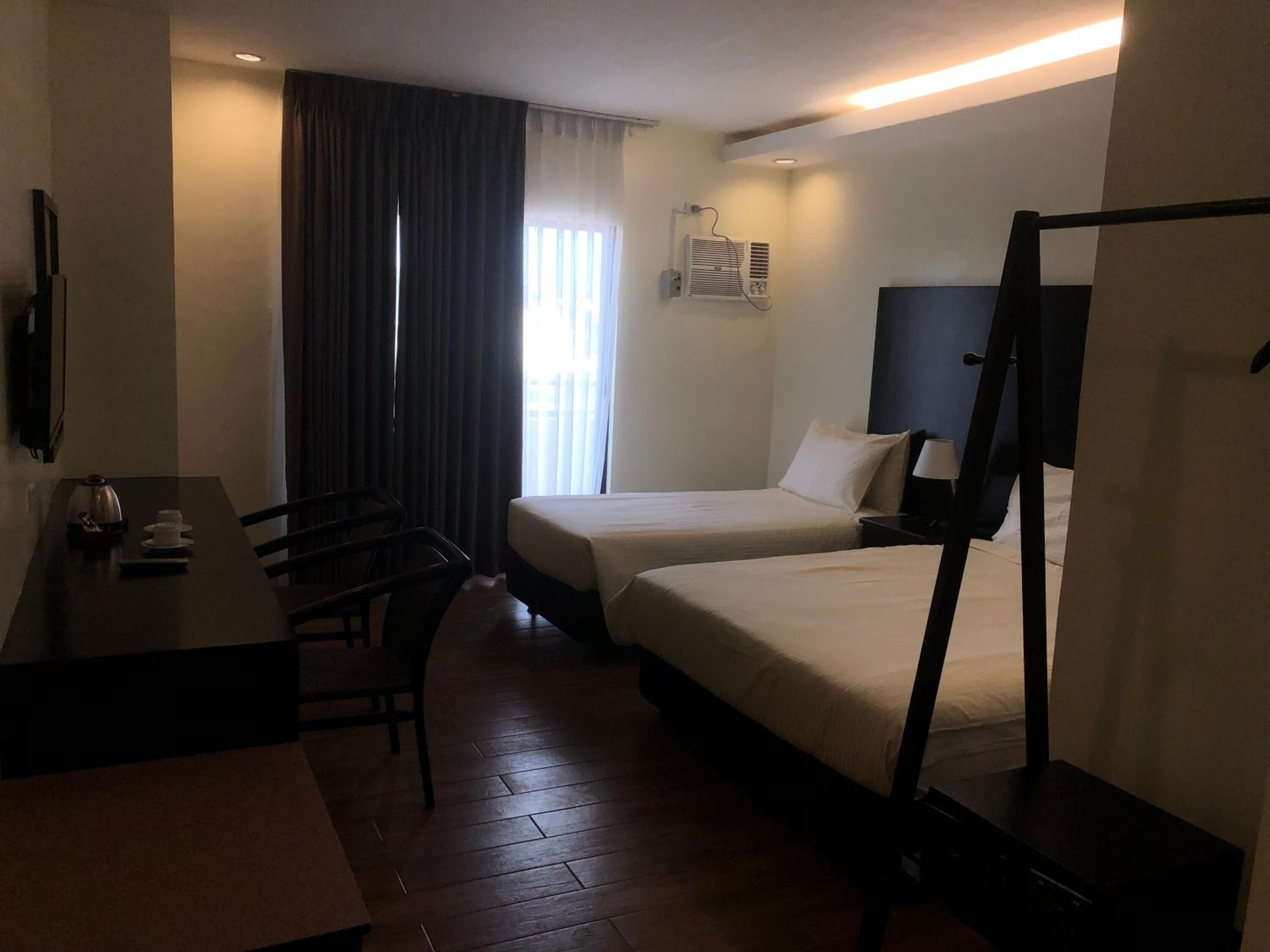 A hotel room with two single beds, a desk with a chair, a TV, and an air conditioning unit.