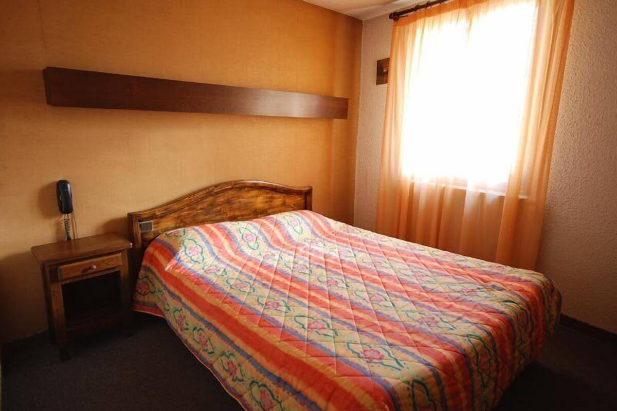 Ein Hotelzimmer mit hölzernem Kopfteil, einer farbenfrohen Bettdecke, einem Nachttisch mit Lampe und einem Vorhangfenster.