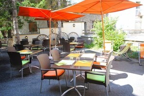 Un café al aire libre con mesas y sillas, algunas con sombrillas naranjas.