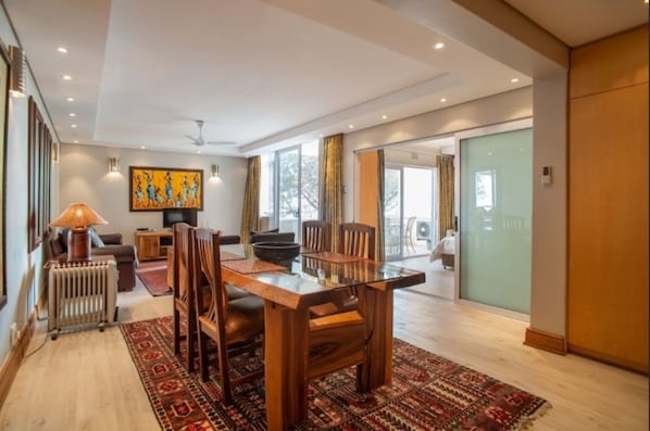 A spacious living area with a wooden dining table, comfortable seating, a radiator, and a large window with curtains.