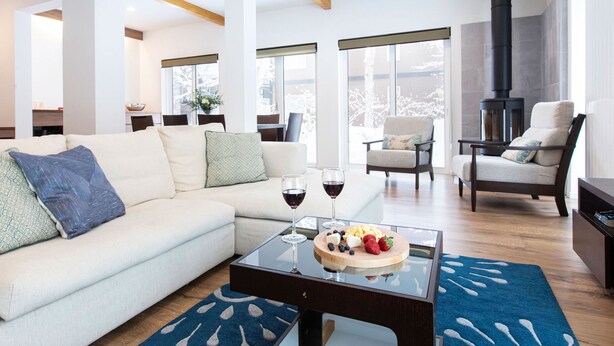 A modern living room with a sofa, chairs, a glass coffee table with fruit, and a fireplace. - Ajisai Chalet  (Kutchan)