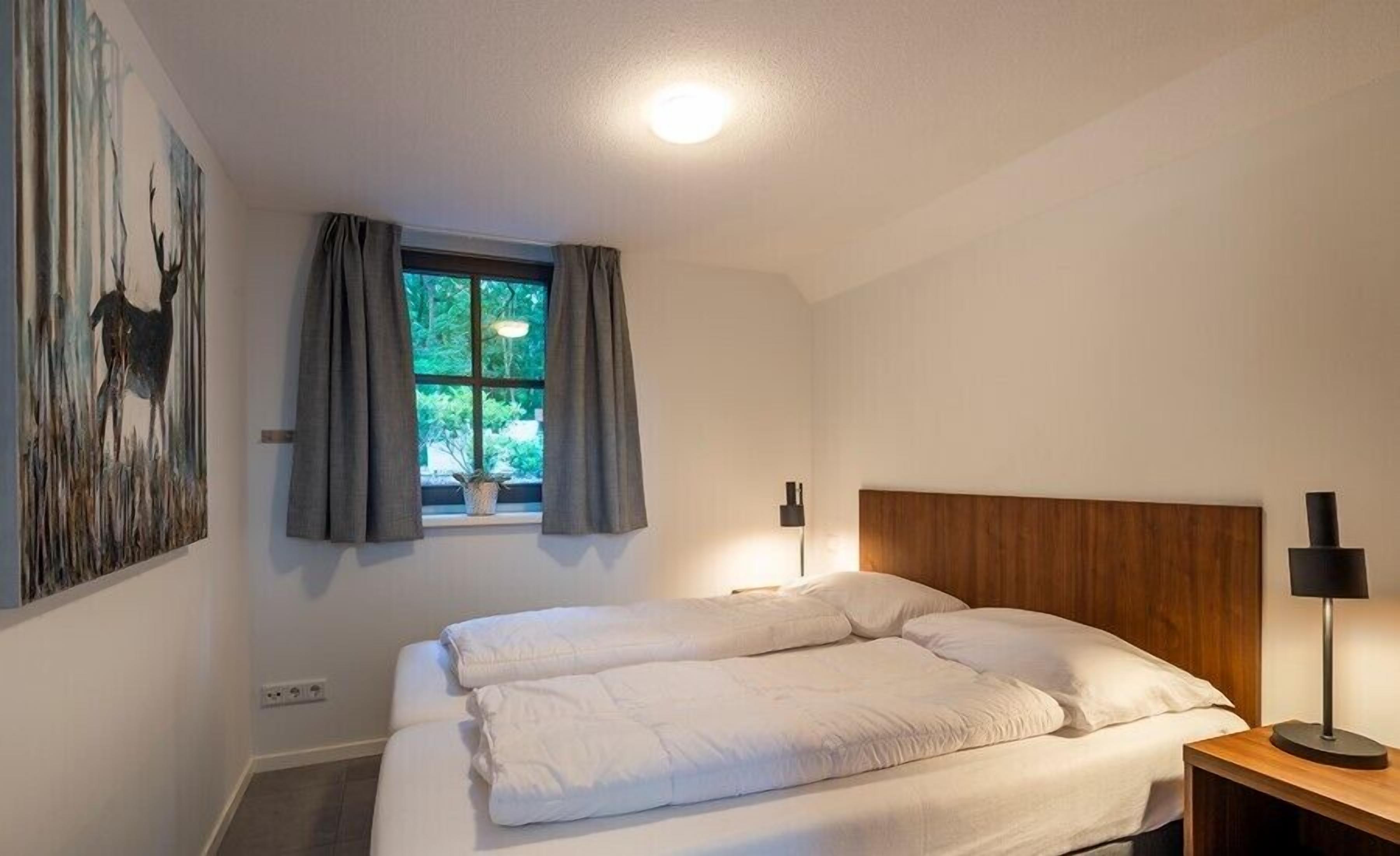 A bedroom with a bed, a window with curtains, a wall-mounted lamp, and a painting.