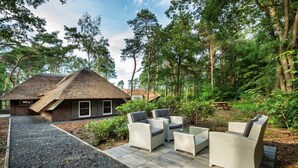 Une maison avec un toit de chaume, entourée d’une forêt. On y trouve une terrasse avec du mobilier d’extérieur.