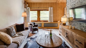Ein gemütliches Zimmer mit Holzbalkendecke, Steinwand, großem Fenster, einer Couch, einem Tisch mit Gläsern und einer Flasche sowie einem Fernseher an der Wand.