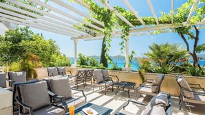 Un espace extérieur aménagé avec des chaises et des tables, une pergola couverte de plantes grimpantes et une vue sur la mer.