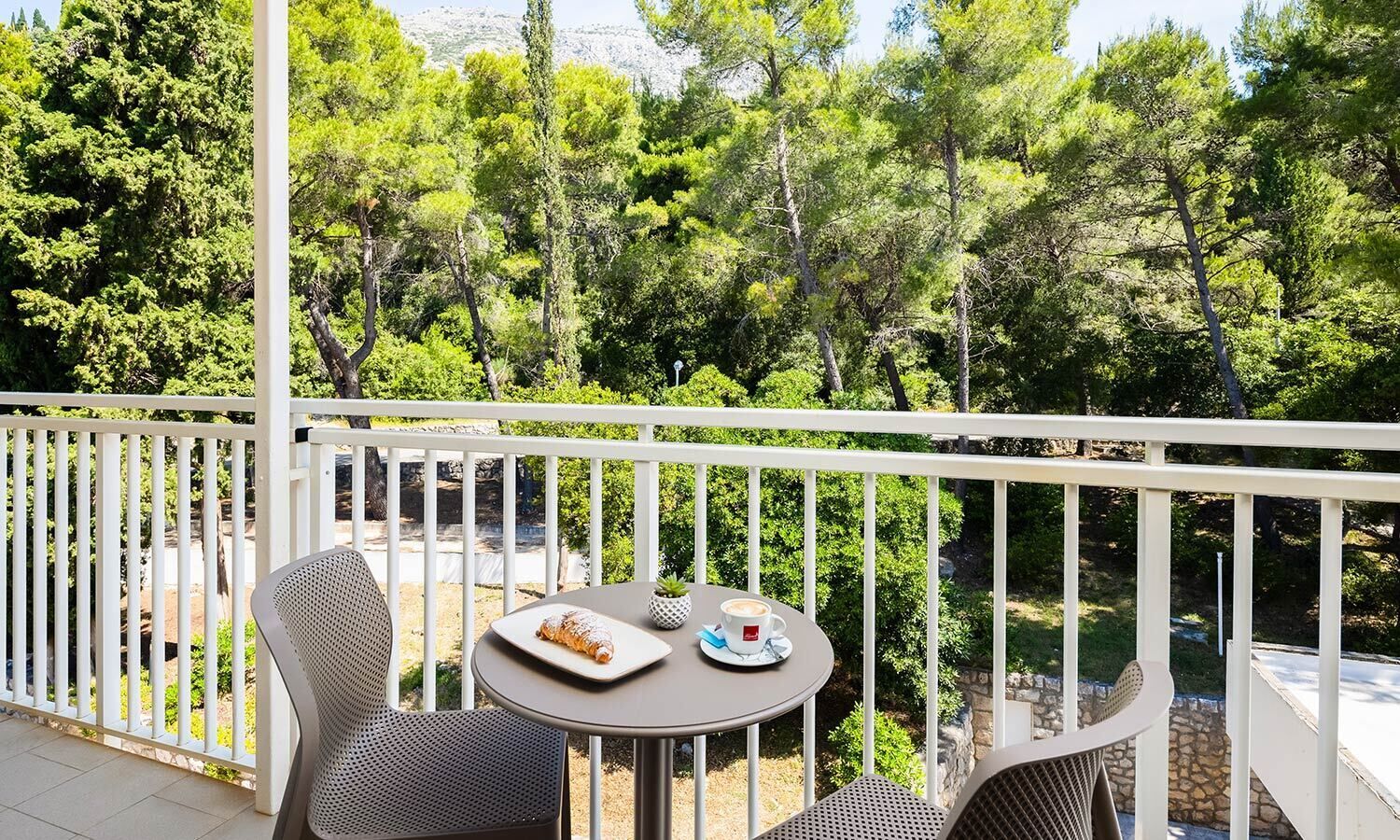 Un balcón con una mesa dispuesta para dos personas, rodeado de barandillas blancas, con vistas a una zona boscosa.