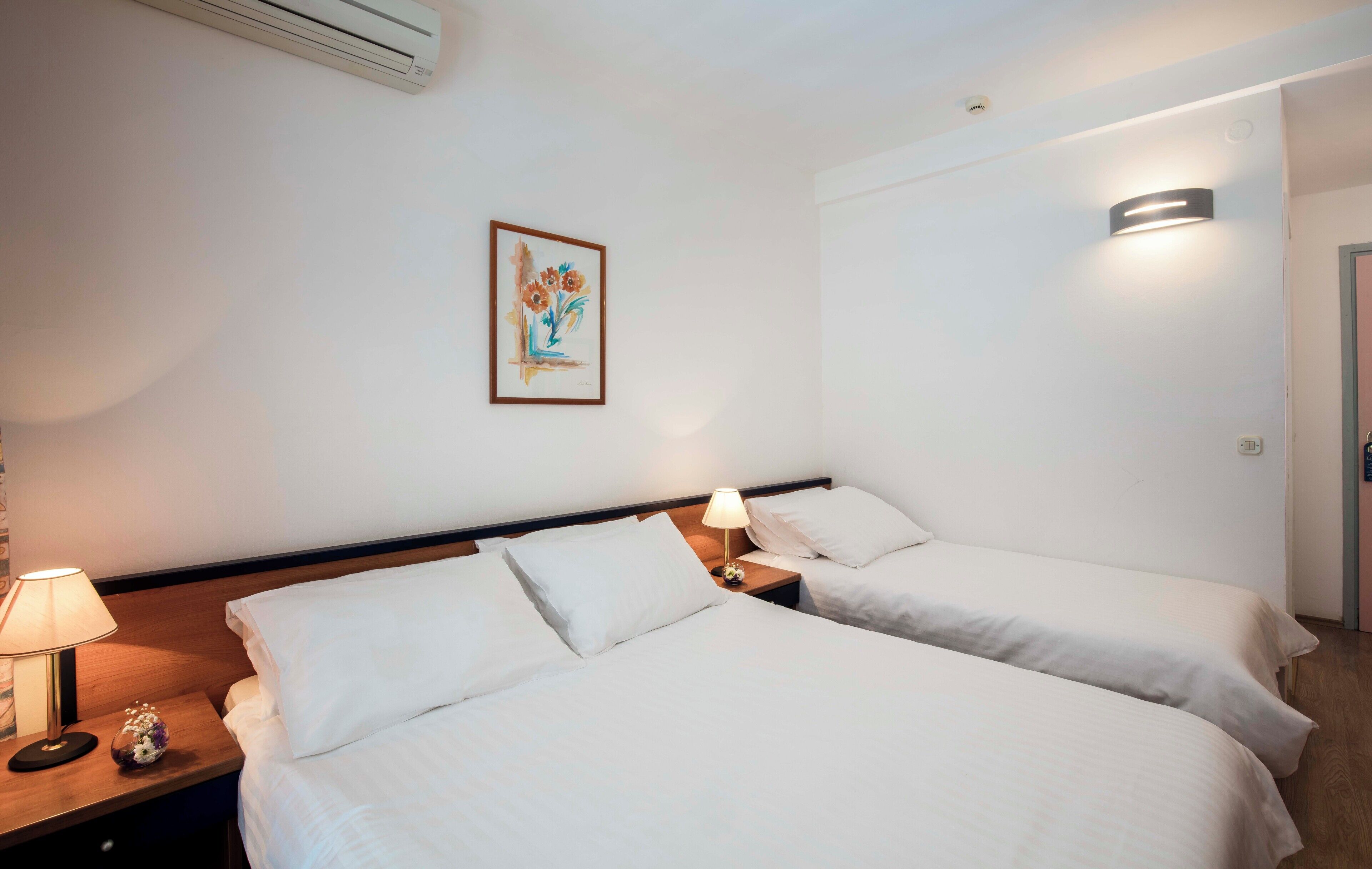 Habitación de hotel con dos camas individuales, una mesita de noche con lámpara, una obra de arte floral enmarcada en la pared y una lámpara de pared.