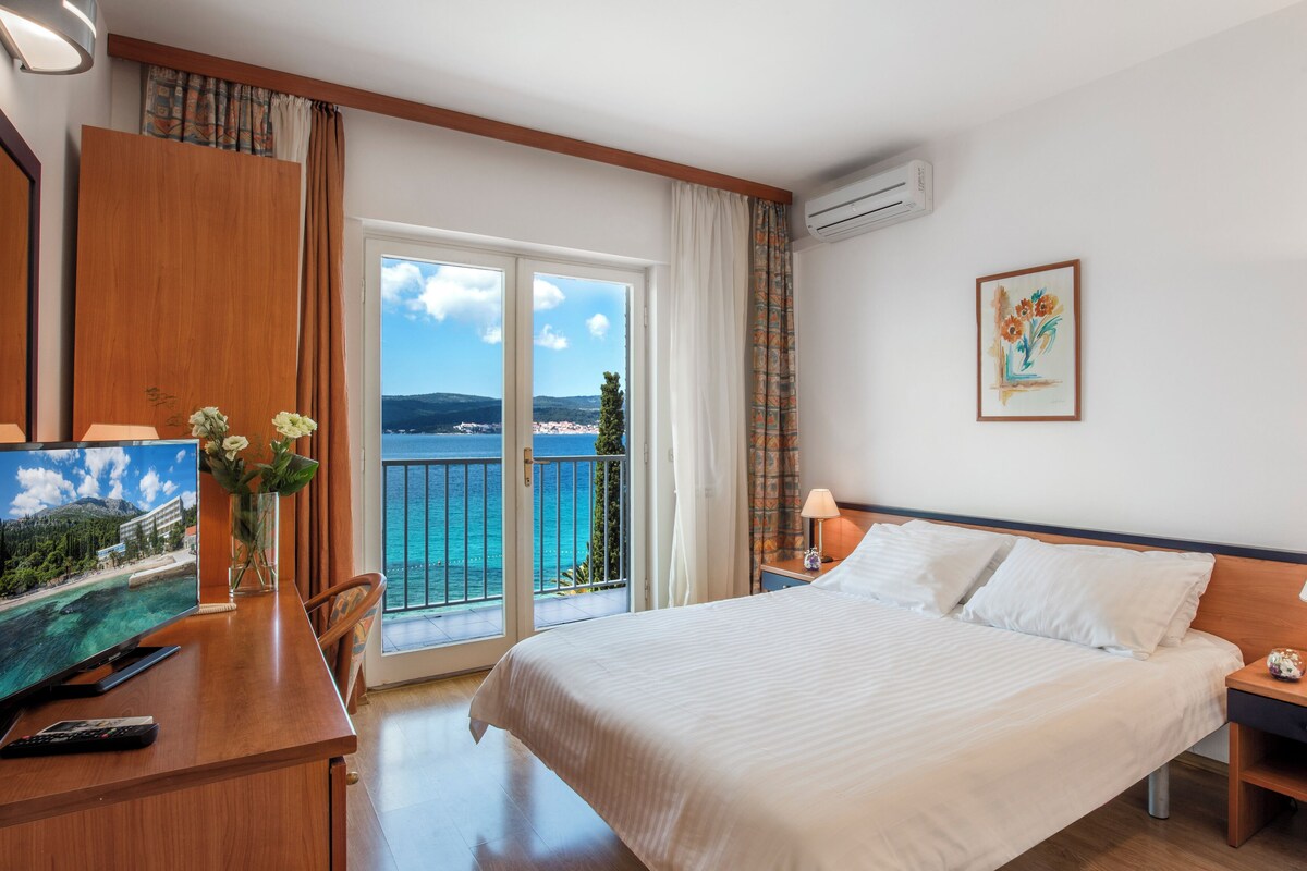 A hotel room with a bed, a desk with a TV, a chair, a nightstand with a lamp, and a painting on the wall.