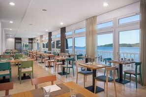 Un restaurante con varias mesas y sillas, amplias ventanas con cortinas y vistas al mar.