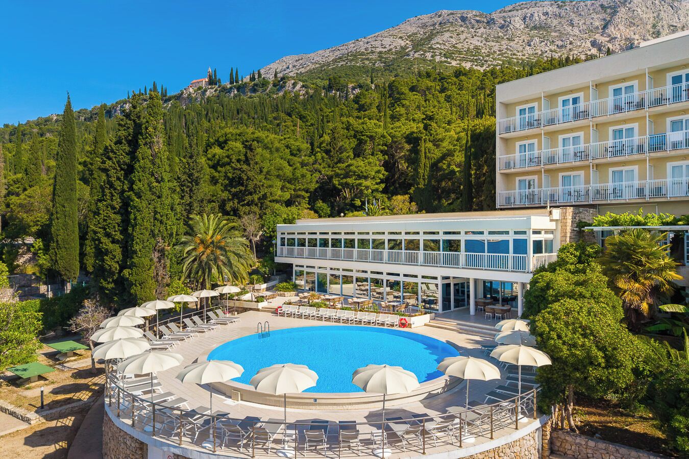 A hotel with a pool surrounded by sun loungers and umbrellas, overlooking a forested hillside.