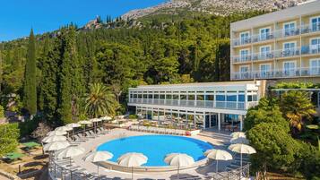A hotel with a pool surrounded by sun loungers and umbrellas, overlooking a forested hillside.
