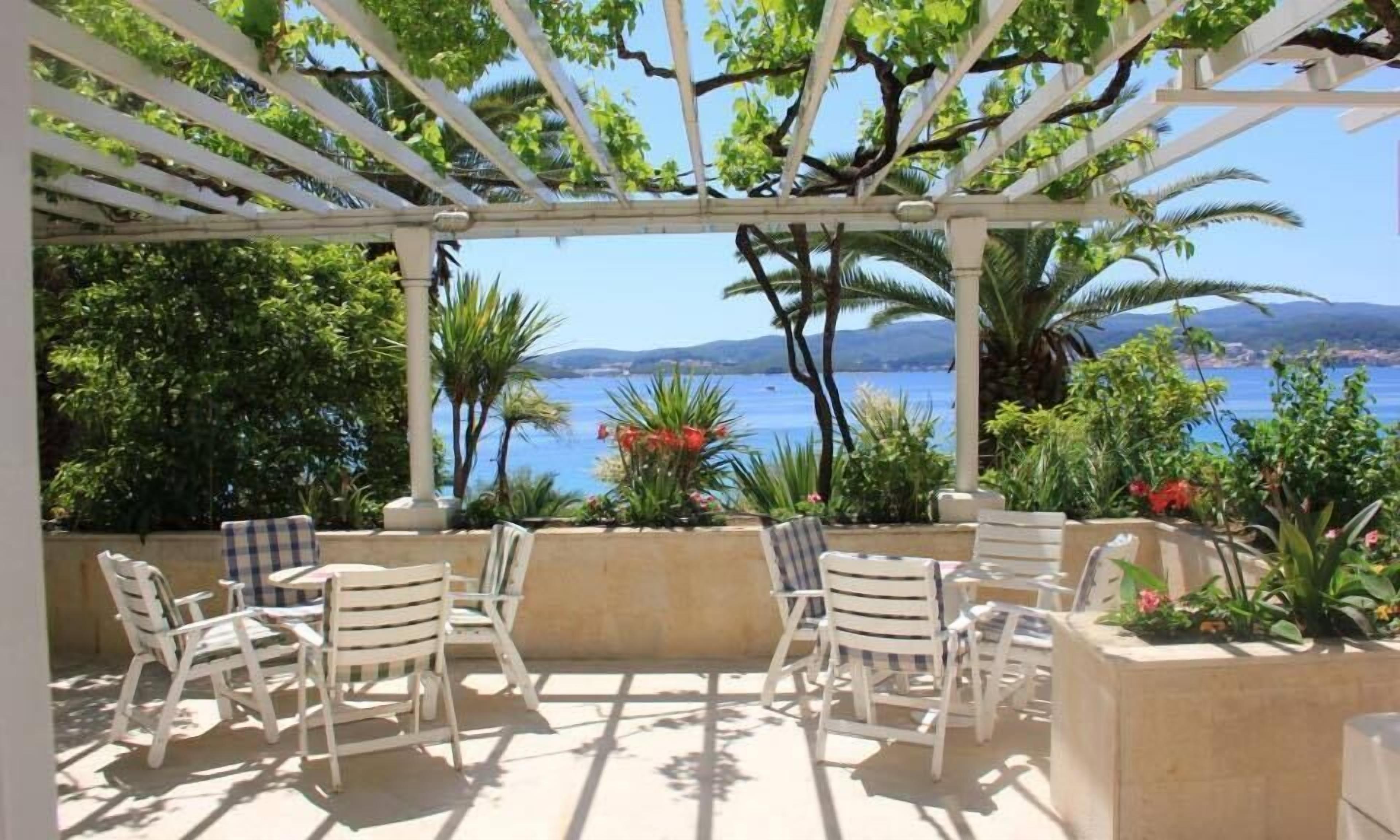 A covered outdoor seating area with white chairs and tables, overlooking a body of water and greenery.