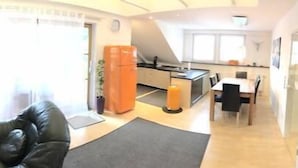 Ein modernes Wohnzimmer mit einer schwarzen Ledersofa, einem Glastisch und einem Fernseher auf einem weißen Schrank. Es gibt eine Küchenzeile mit einem Kühlschrank und einem Esstisch mit Stühlen.