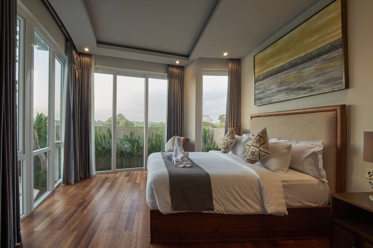 A spacious bedroom with a large bed, a view through floor-to-ceiling windows, and a painting on the wall.