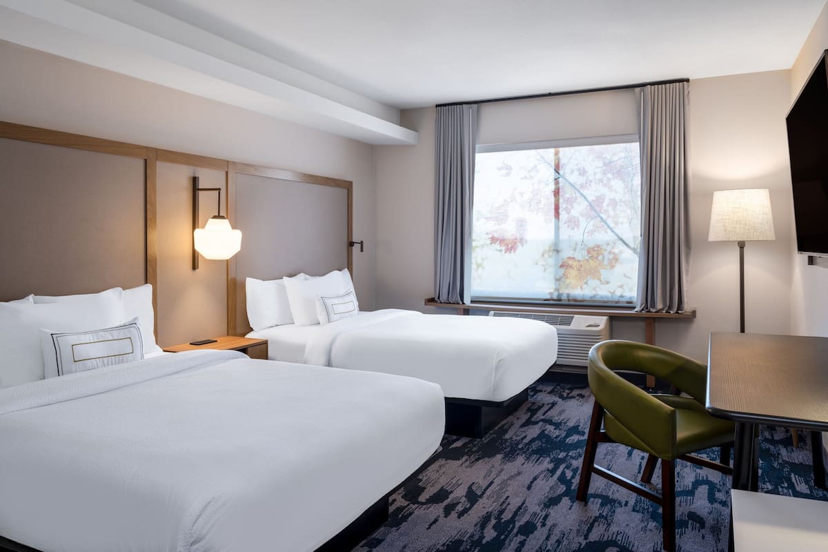 Habitación de hotel con dos camas, un escritorio, una silla, televisión y ventana con cortinas.