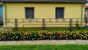 A yellow single-story house with a brown tiled roof, two windows, and a small garden with yellow flowers. - Szigeti Ház, Szécsisziget (Deák Ferenc utca)