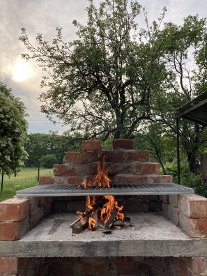 An outdoor brick grill with flames, surrounded by trees and a clear sky. - Szigeti Ház, Szécsisziget (Deák Ferenc utca)
