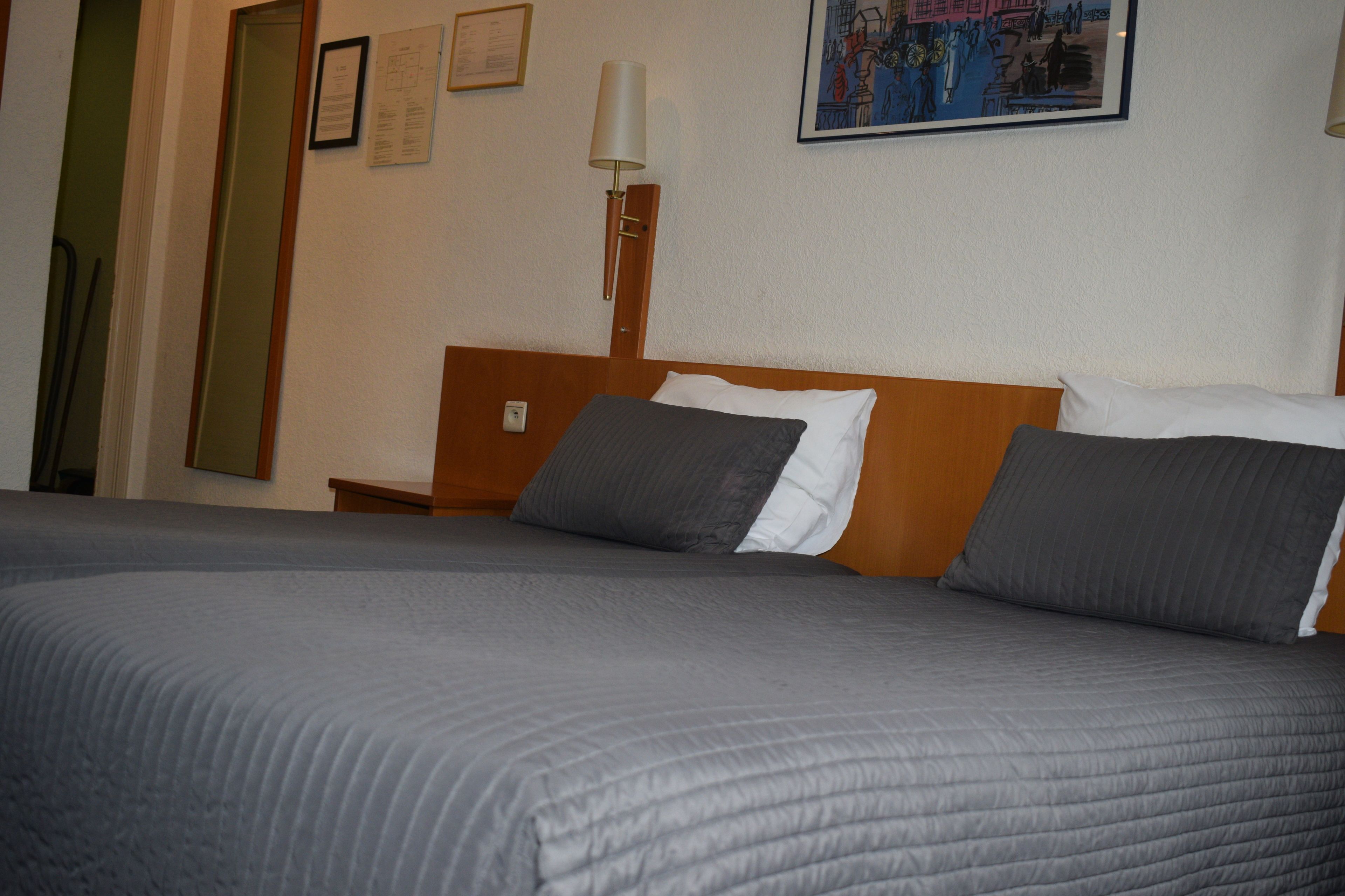 A neatly made bed with a grey quilt, a wooden headboard, and a wall-mounted lamp.