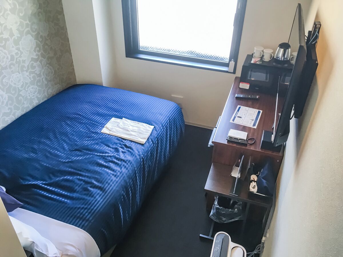 Et lille hotelværelse med en enkeltseng, et skrivebord med computer, en stol og et vindue.