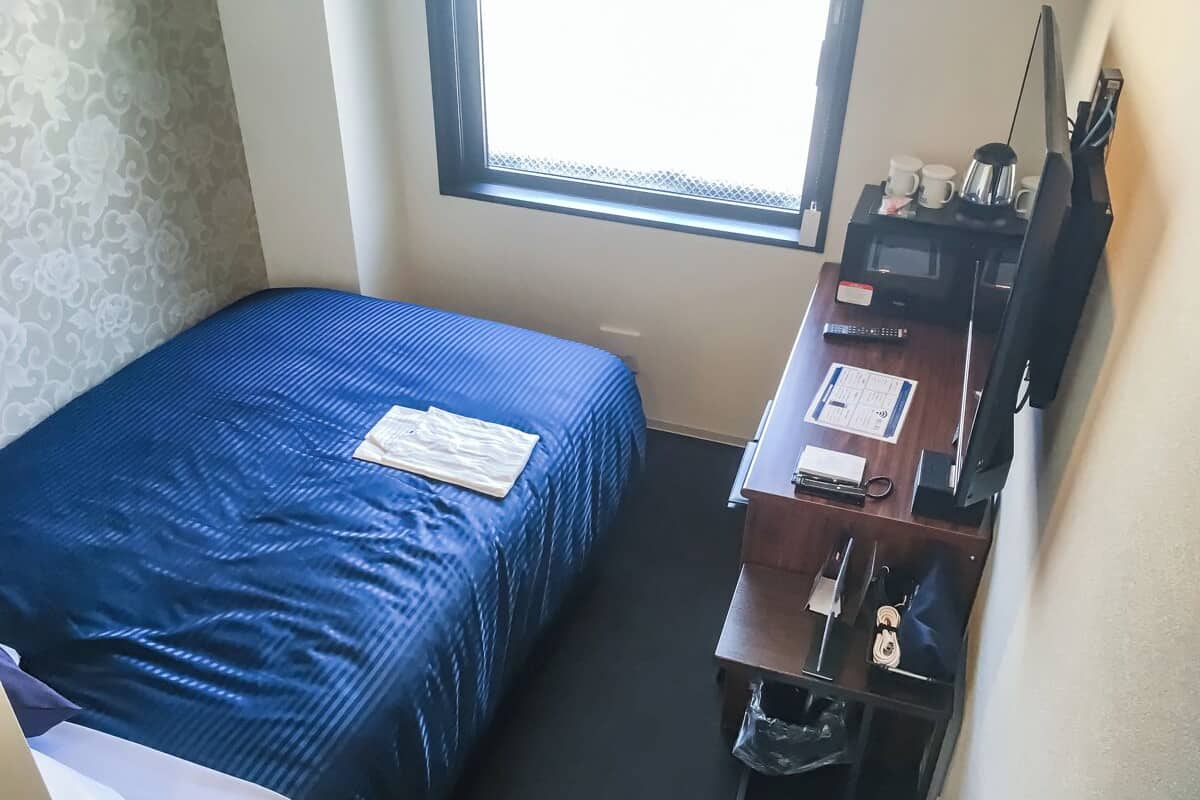 Quarto de hotel pequeno com uma cama de solteiro, uma escrivaninha com computador, uma cadeira e uma janela.